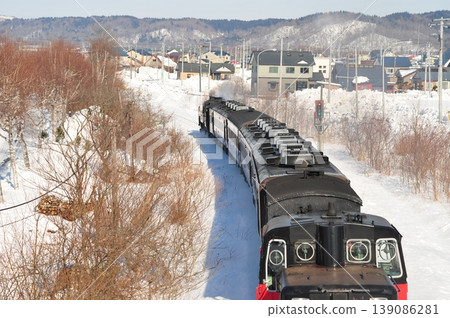 一輛蒸汽機車行駛在北海道白雪皚皚的景色和當地城鎮風光中。 139086281