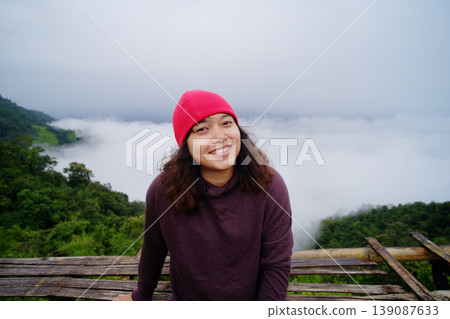 Smiling tourist Asian young woman wearing red hat and sitting on wooden bench on peak of mountains and mist in the morning of Thailand Smiling tourist Asian young woman wearing red hat and sitting on wooden bench on peak of mountains and mist in the morning of Thailand 139087633