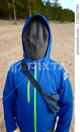 Unique figure stands alone on a beach wearing a blue jacket and covered face 139087990