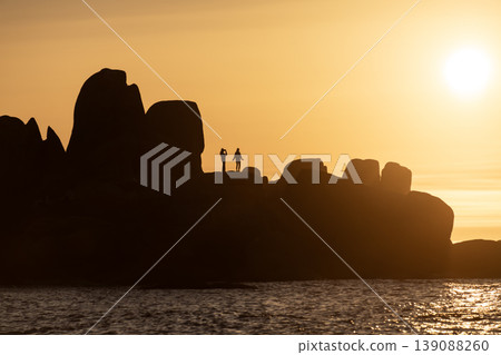 Silhouette of rocky coastline with people walking during dramatic golden sunset over calm sea 139088260