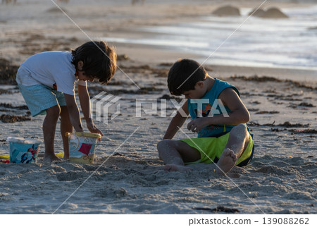 Two children playing with colorful sand toys on beach during warm sunset by ocean shore Two children playing with colorful sand toys on beach during warm sunset by ocean shore 139088262
