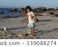 Child standing on beach with rocks and ocean background holding bucket during summer day 139088274