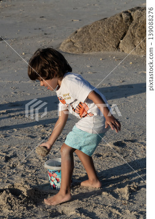 Young child playing with sand on beach bending down near rocks during warm sunny day Young child playing with sand on beach bending down near rocks during warm sunny day 139088276