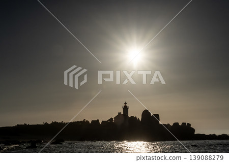 Sunset silhouette of lighthouse and rocky coastline with bright sun reflecting over calm sea Sunset silhouette of lighthouse and rocky coastline with bright sun reflecting over calm sea 139088279