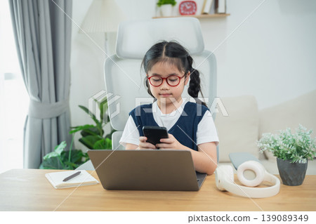 Young girl with glasses using smartphone while sitting at desk with laptop and stationery in modern home office setting 139089349