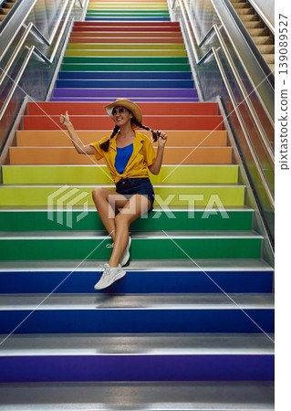 Smiling woman sits on steps of an underground passage painted in LGBT Rainbow colors and shows thumbs up Smiling woman sits on steps of an underground passage painted in LGBT Rainbow colors and shows thumbs up 139089527
