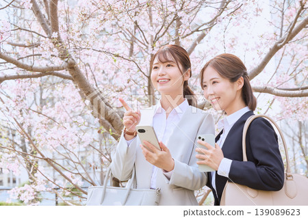 Two young female businesswomen in suits enjoying cherry blossom viewing while holding their smartphones under the cherry trees. Two young female businesswomen in suits enjoying cherry blossom viewing while holding their smartphones under the cherry trees. 139089623