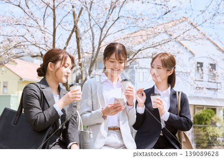 三位年輕的職業婦女在春日午休時間，坐在櫻花樹下，一邊喝著咖啡，一邊看著智慧型手機聊天。 139089628