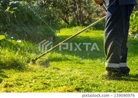 Gardener using string trimmer to cut grass in backyard. Man maintaining lawn with edge trimmer. Landscaping and yard work concept. Home garden service and seasonal turf care professional. Gardener using string trimmer to cut grass in backyard. Man maintaining lawn with edge trimmer. Landscaping and yard work concept. Home garden service and seasonal turf care professional. 139090976