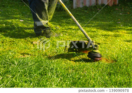 Man mowing grass with string trimmer. Gardener trimming lawn in summer. Professional landscaping and yard maintenance. Powerful tool for horticulture, backyard improvement and garden care. Man mowing grass with string trimmer. Gardener trimming lawn in summer. Professional landscaping and yard maintenance. Powerful tool for horticulture, backyard improvement and garden care. 139090978