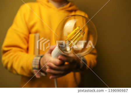 Person in yellow hoodie holding a large vintage Edison light bulb. Concept of creative idea, innovation, and inspiration. Close-up of a decorative lamp in hands with a blurred background 139090987