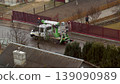 Aerial view of a utility service truck with a cherry picker platform parked on a suburban street. Workers in helmets performing maintenance near a residential fence 139090989