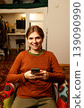 Happy young woman sitting in a colorful armchair and using a smartphone at home. Smiling female scrolling social media or texting on mobile phone in a cozy interior. Gen z girl 139090990