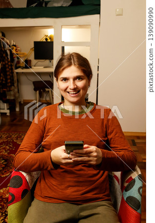 Happy young woman sitting in a colorful armchair and using a smartphone at home. Smiling female scrolling social media or texting on mobile phone in a cozy interior. Gen z girl 139090990