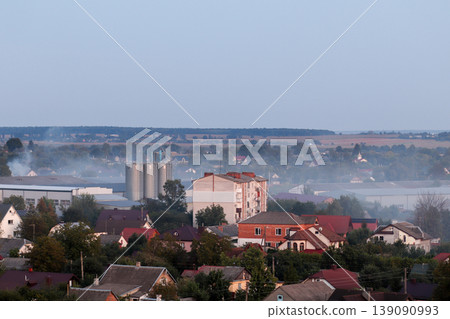 Air pollution from industrial factory pipes over a small town. Concept of environmental issues, smog, and carbon emissions affecting residential areas. Grain elevator 139090993