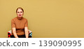Middle-aged woman sitting in a colorful armchair against a solid yellow background with large copy space. Portrait of a calm female in a brown t-shirt. Minimalist studio photography for advertising 139090995