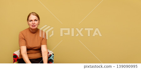 Middle-aged woman sitting in a colorful armchair against a solid yellow background with large copy space. Portrait of a calm female in a brown t-shirt. Minimalist studio photography for advertising 139090995