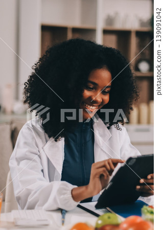 Young nutritionist showing bell peppers during an online video call consulting about healthy eating, sitting at home at a table with vegetables, laptop and measuring tape 139091042