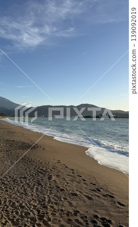 Beach landscape with gentle waves meeting sandy shore during sunset, with mountains in background Beach landscape with gentle waves meeting sandy shore during sunset, with mountains in background 139092019