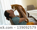A brunette caucasian woman kisses and plays with a ginger domestic cat while sitting in a kitchen 139092579