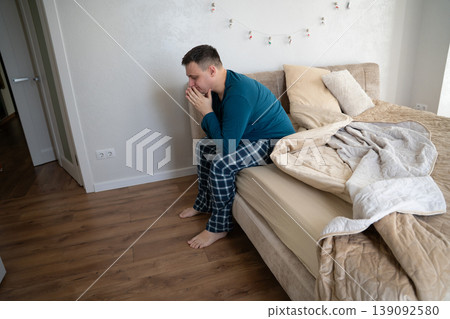 35 year old caucasian man sits on his bed in his pajamas in the bedroom in the morning. Waking up after a night's sleep and routine, lifestyle concept 139092580