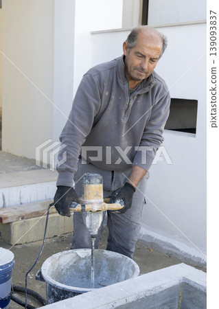 Construction worker mixing mortar with electric mixer in bucket during exterior renovation Construction worker mixing mortar with electric mixer in bucket during exterior renovation 139093837