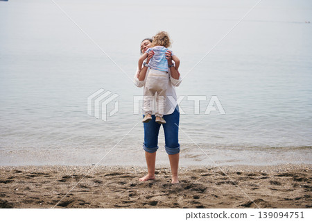 Mother Hugging Young Child on Beach Shore During Calm Day, Family Bonding Moment 139094751