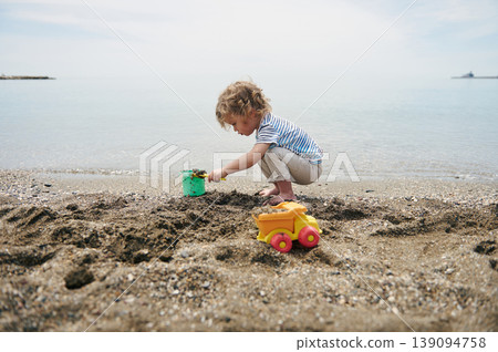 Little Boy Playing in Sand on Quiet Beach With Toy Truck and Bucket at Seaside Little Boy Playing in Sand on Quiet Beach With Toy Truck and Bucket at Seaside 139094758