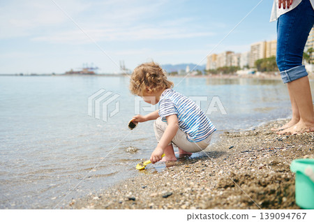 Child Playing on Pebble Beach With Parent Standing Nearby on Sunny Coastline 139094767