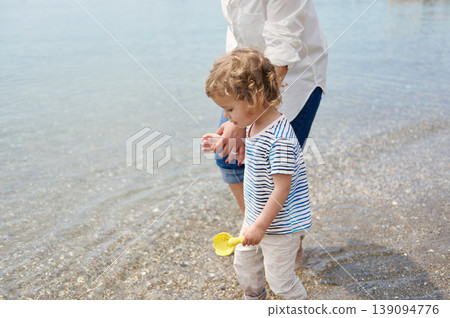 Toddler Boy Playing On Shallow Beach With Parent Holding Hand During Sunny Summer Day 139094776