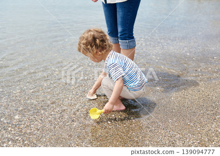 Toddler Playing With Seashells on Pebble Beach Beside Parent at Shallow Shore 139094777