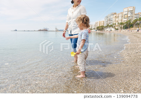 Mother and Young Child Walking on Sunny Beach Shore With City Skyline 139094778