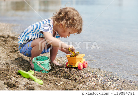Curious Toddler Playing With Sand Toys On A Sunny Beach Shore 139094781