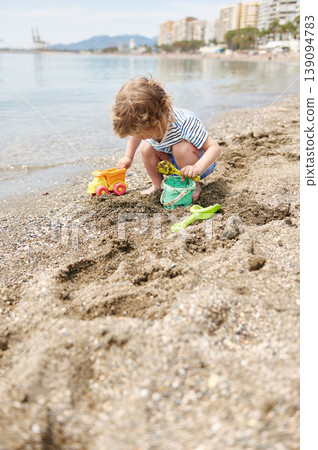 Toddler Playing With Sand Toys On Sunny Urban Beach Near Waterfront Buildings 139094783