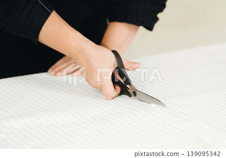 A woman seamstress cuts a white fabric on the table. A woman seamstress cuts a white fabric on the table. 139095342