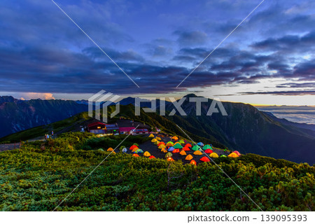 Dawn at Mt. Chogatake in the Northern Alps: Hikers waiting for the sunrise, and the illuminated mountain huts of Mt. Jonen. Dawn at Mt. Chogatake in the Northern Alps: Hikers waiting for the sunrise, and the illuminated mountain huts of Mt. Jonen. 139095393