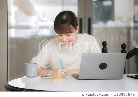 Happy smiling asian woman working at home enjoying studying online business course using modern technology writing notes on paper by computer sitting at white round table inside room Happy smiling asian woman working at home enjoying studying online business course using modern technology writing notes on paper by computer sitting at white round table inside room 139095679