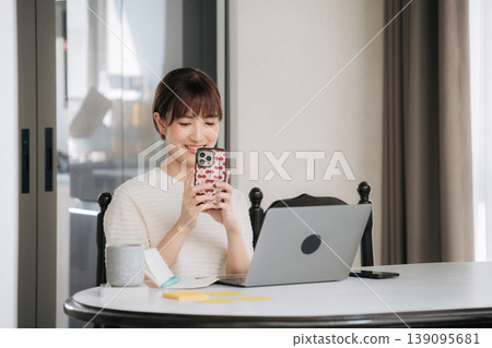 Happy asian woman enjoying working from home using modern technology while studying online for business management looking at smartphone screen smiling brightly next to laptop computer Happy asian woman enjoying working from home using modern technology while studying online for business management looking at smartphone screen smiling brightly next to laptop computer 139095681