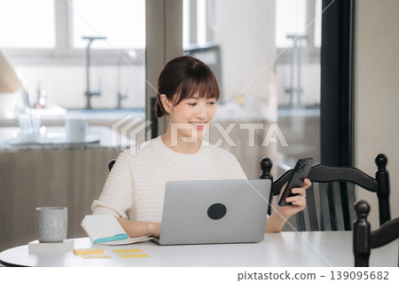 Happy asian woman working at home using technology for studying and online business smiling brightly while looking at smartphone sitting by laptop Happy asian woman working at home using technology for studying and online business smiling brightly while looking at smartphone sitting by laptop 139095682