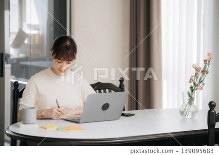 Focused young asian woman happily working online business from home using modern technology while calmly studying and writing important notes at dining table 139095683