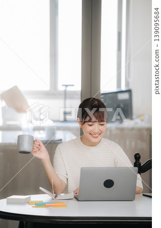 Happy asian woman working from home using technology for studying online business while drinking coffee holding mug smiling feeling relaxed and joyful looking at laptop screen sitting by window 139095684