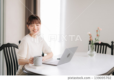 Happy smiling asian woman sitting comfortably at home while working on modern computer technology and studying for an online business class drinking coffee Happy smiling asian woman sitting comfortably at home while working on modern computer technology and studying for an online business class drinking coffee 139095702