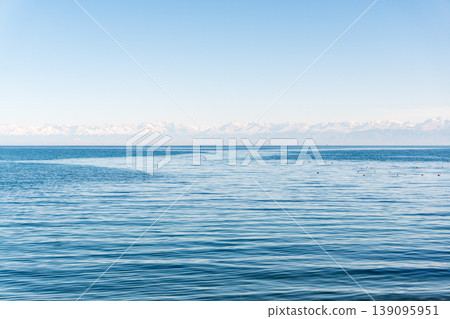 Wide landscape of blue water and distant snow-capped mountains. Alpine lake view under a clear sky with a flock of birds on the surface. Nature background with copy space for text Wide landscape of blue water and distant snow-capped mountains. Alpine lake view under a clear sky with a flock of birds on the surface. Nature background with copy space for text 139095951