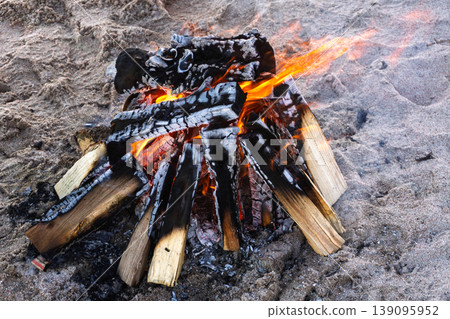 Burning campfire on a sandy beach. Bright orange flames and charred wood logs. Outdoor camping and survival concept Burning campfire on a sandy beach. Bright orange flames and charred wood logs. Outdoor camping and survival concept 139095952