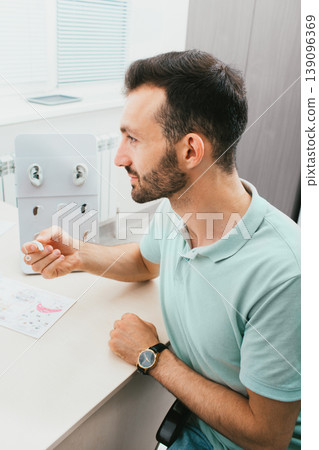 Close-up portrait of an adult male with hearing impairment during the selection of hearing aids in a hearing clinic. Close-up portrait of an adult male with hearing impairment during the selection of hearing aids in a hearing clinic. 139096369