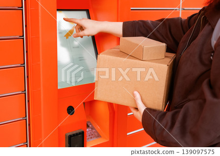 Woman holding cardboard boxes while using the touchscreen of an orange automated parcel locker for delivery or pickup. E-commerce and logistics concept. Woman holding cardboard boxes while using the touchscreen of an orange automated parcel locker for delivery or pickup. E-commerce and logistics concept. 139097575