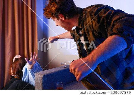 Aggressive drunk man holding a bottle preparing to punch his scared wife hiding behind the sofa. Concept of domestic violence, abuse, alcoholism, anger, and dangerous assault. Aggressive drunk man holding a bottle preparing to punch his scared wife hiding behind the sofa. Concept of domestic violence, abuse, alcoholism, anger, and dangerous assault. 139097579