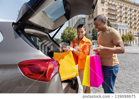 Happy young Black couple loading colorful shopping bags into car trunk Happy young Black couple loading colorful shopping bags into car trunk 139098675
