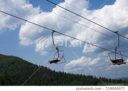 Empty chairlift chairs moving against a blue sky over green mountains Empty chairlift chairs moving against a blue sky over green mountains 139098971