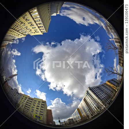 Looking straight up into a bright blue sky filled with white clouds framed by tall urban apartment buildings. The scene captures daily life in a lively city setting. 139100473